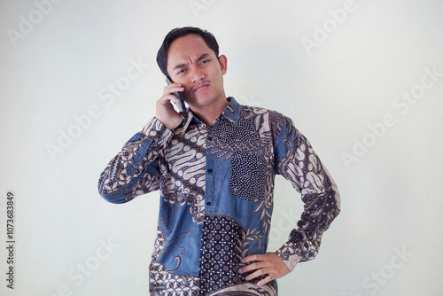 Thoughtful young asian man in a batik shirt holding a phone to his ear, looking puzzled and deep in thought against a plain background.
