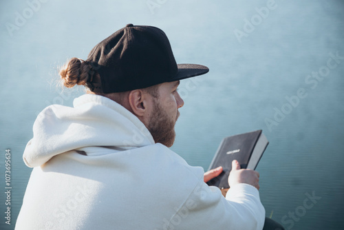 A young man reads the Bible while sitting by the lake at sunset.