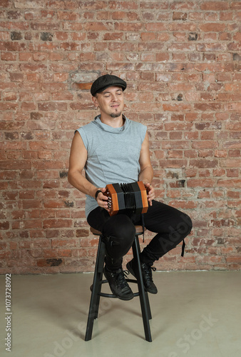 Male musician seated on stool holding concertina, against brick wall background photo