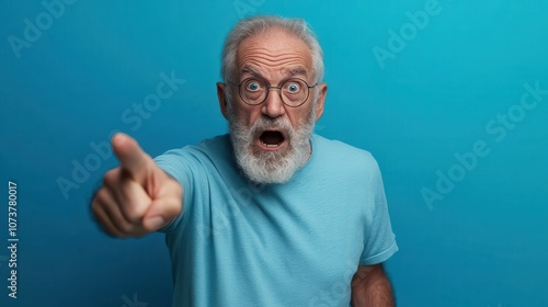 An elderly man with a surprised expression is seen pointing outward while wearing a light blue t-shirt against a vibrant blue background.