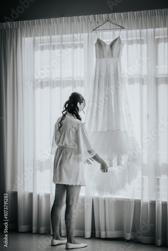 Free Download A bride in a white robe gently touches her wedding dress, hanging by a window with soft natural light filtering through sheer curtains in a serene setting.. - GFXLoad