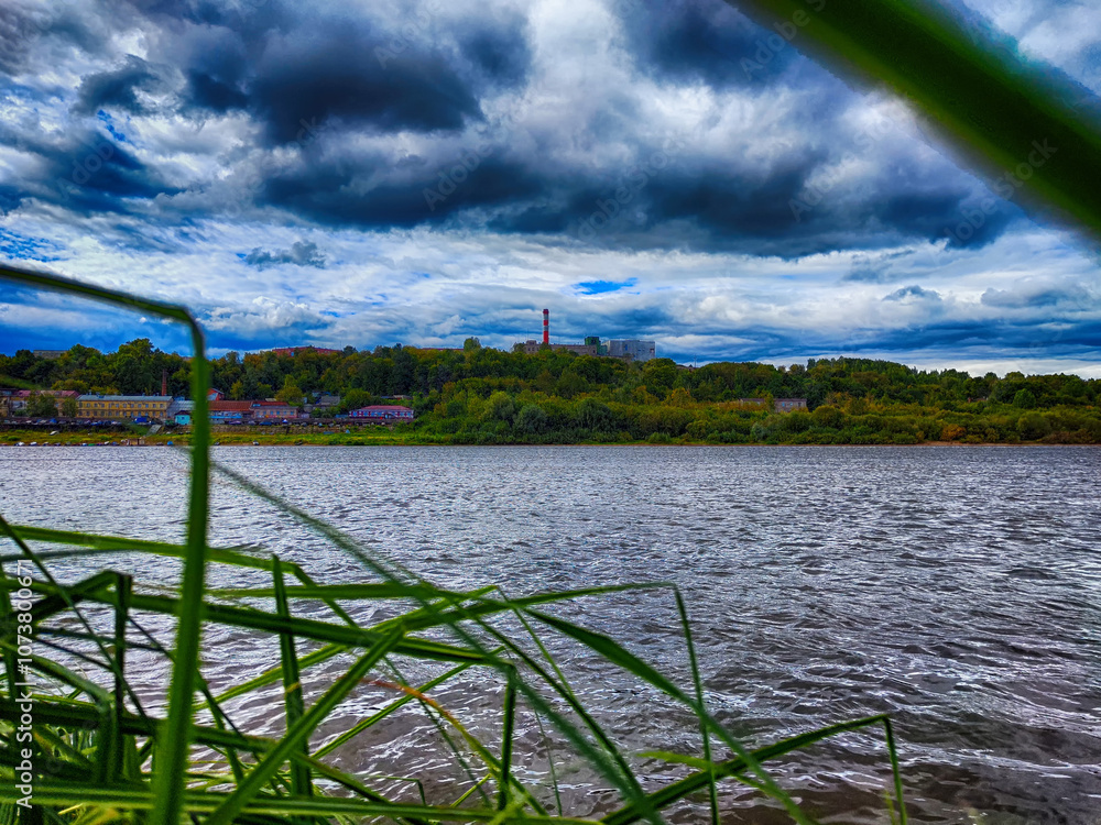 Cloudy sky over a river with tall grasses in the foreground and a distant landscape along the shoreline Cloudy sky over a river with tall grasses in the foreground and a distant landscape along the shoreline