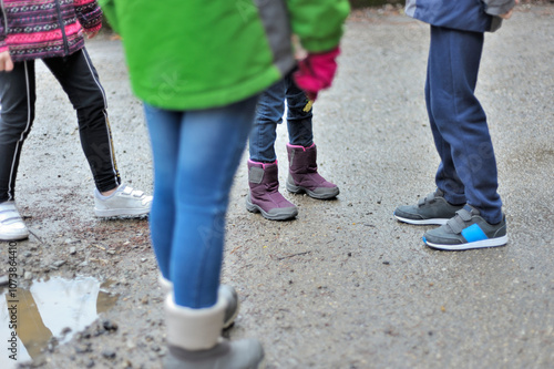 Horizontal photograph of 4 unrecognizable children playing and gathered in the street next to a small puddle of water, wearing sports shirts, wellies and rain and cold clothes. Selective focus