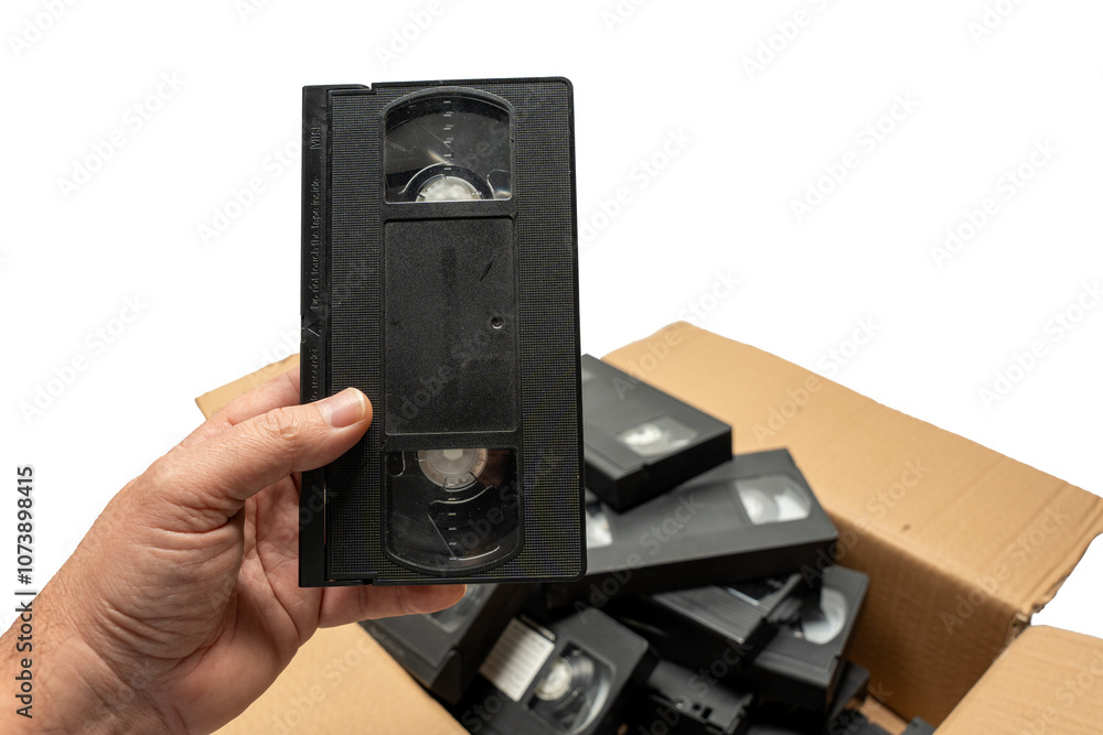 Obraz premium A person is holding a black VHS tape in a box full of other black VHS tapes. The box is filled with various tapes, some of which are in different sizes and shapes