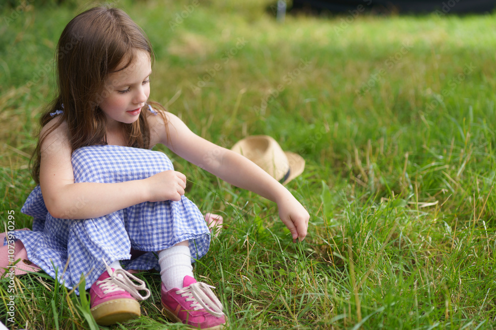 Fototapeta premium Little cute girl in a sundress and a straw hat sits on the lawn. The concept of children's health, education, fun
