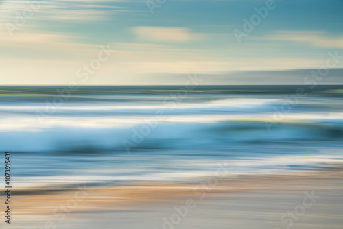 Light airy abstract seascape. Wide sandy beach, sea waves, silhouette of mountains, and cloudy sky in the background, motion blur