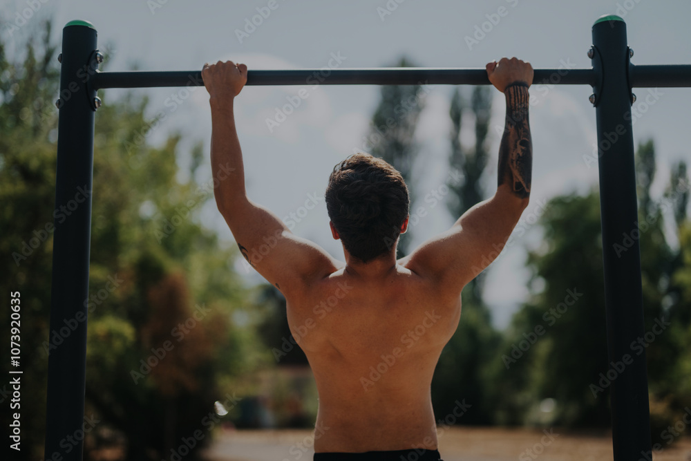 Naklejka premium Rear view of a shirtless young man performing pull-ups on a bar in a park, showcasing fitness, strength, and a healthy outdoor lifestyle.