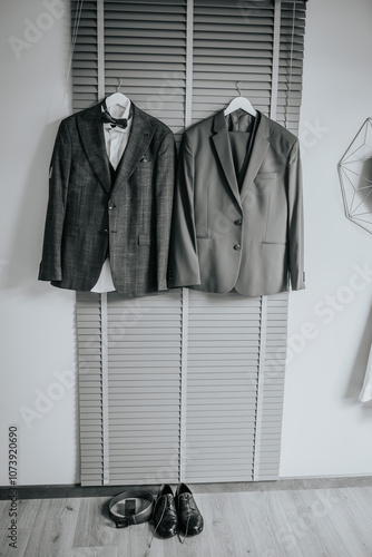 Two suit jackets on hangers with a pair of dress shoes and a belt on the floor, representing formalwear preparation for a wedding or event.. photo