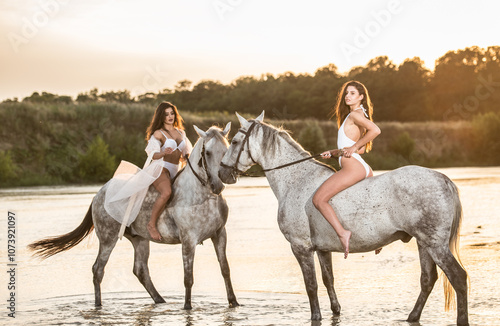 Beautiful brunettes woman with long hair ,white bikini and white shirt rides  on a white horse in the river. Sensual and attractive girls  interacting and rides a horse in the water at sunset