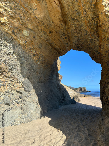 A captivating rock arch formation carved naturally over time, standing on a sandy beach under a bright blue sky.
