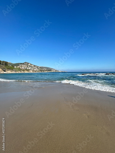 A serene view of a pristine sandy beach stretching out towards the horizon, with gentle waves caressing the shoreline under a brilliant, cloudless blue sky.