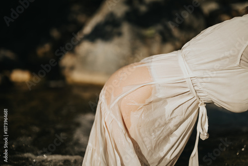 A woman is seen bending over in a white dress near a stream, with sunlight highlighting her wet attire. The scene captures both nature and tranquility.