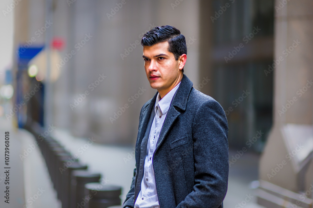 A man in a formal coat stands thoughtfully on a city street, surrounded by blurred buildings and bollards. He has dark hair and an alert expression, suggesting he's deep in thought.
