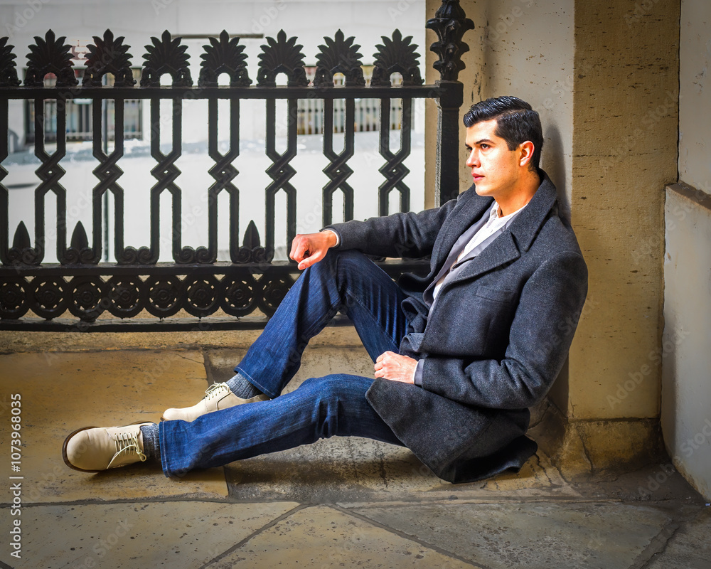 A man in a dark coat and jeans sits casually against a stone wall, with an ornate metal fence in the background. He appears thoughtful, gazing into the distance.