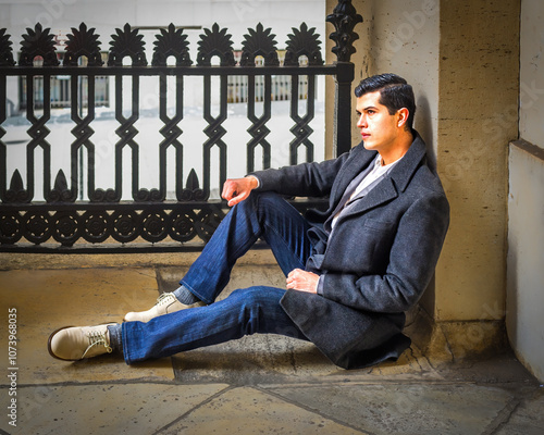 Obraz na płótnie A man in a dark coat and jeans sits casually against a stone wall, with an ornate metal fence in the background. He appears thoughtful, gazing into the distance. w salonie