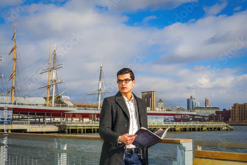 Obraz na płótnie A man dressed in a coat stands by the waterfront, holding an open book. Behind him, several tall ships and a city skyline are visible under a partly cloudy sky. w salonie
