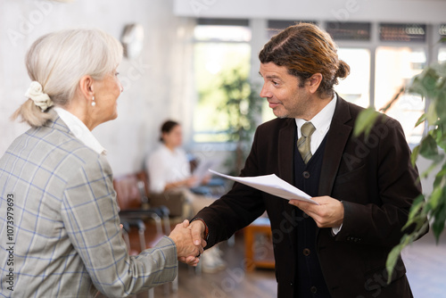 Smiling businessman holding papers and shaking hands with elderly woman as sign of closing deal on meeting in office photo