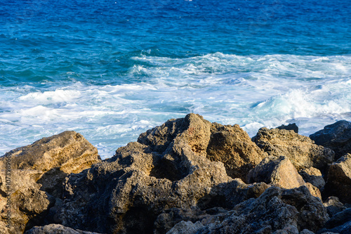 Waves crash against rugged rocks under the bright sun on a serene coastal day.