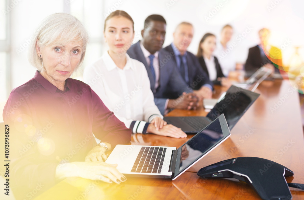 Fototapeta premium Senior businesswoman using laptop during conference in meeting room. Woman sitting at desk with colleaugues.
