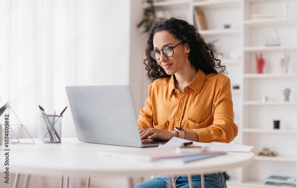 © Prostock-studio - Middle Eastern Lady Using Laptop Working Online Wearing Eyewear Sitting At Workplace In Modern Office. Remote Job, Technology And Career Profession Concept. Side View