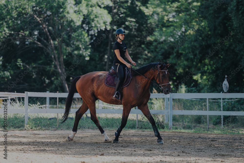 A person rides a brown horse in a fenced outdoor paddock surrounded by green trees. The equestrian is wearing a helmet for safety. The scene is calm and serene, capturing a sunny day. A person rides a brown horse in a fenced outdoor paddock surrounded by green trees. The equestrian is wearing a helmet for safety. The scene is calm and serene, capturing a sunny day.