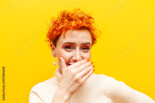 young red-haired woman with curly hair ridicules and mocks on yellow isolated background, girl with beautiful make-up is shy and covers her smile with her hand photo