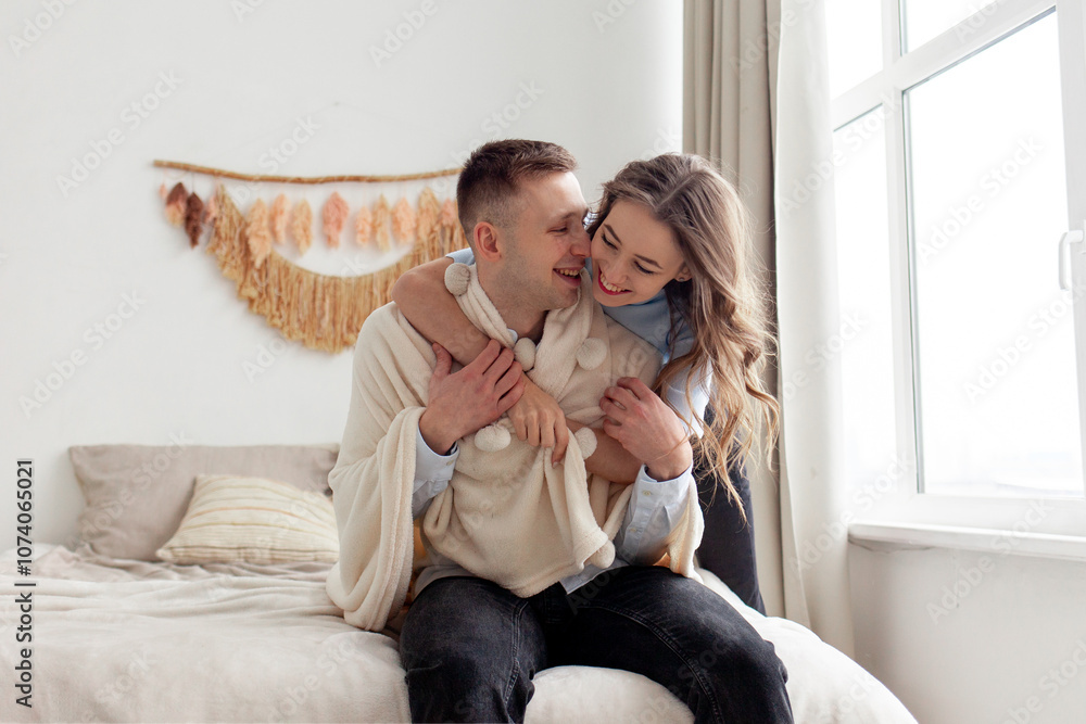 young happy couple in love hugging on the bed at home and smiling, the woman covers the man with a warm soft blanket and takes care young happy couple in love hugging on the bed at home and smiling, the woman covers the man with a warm soft blanket and takes care