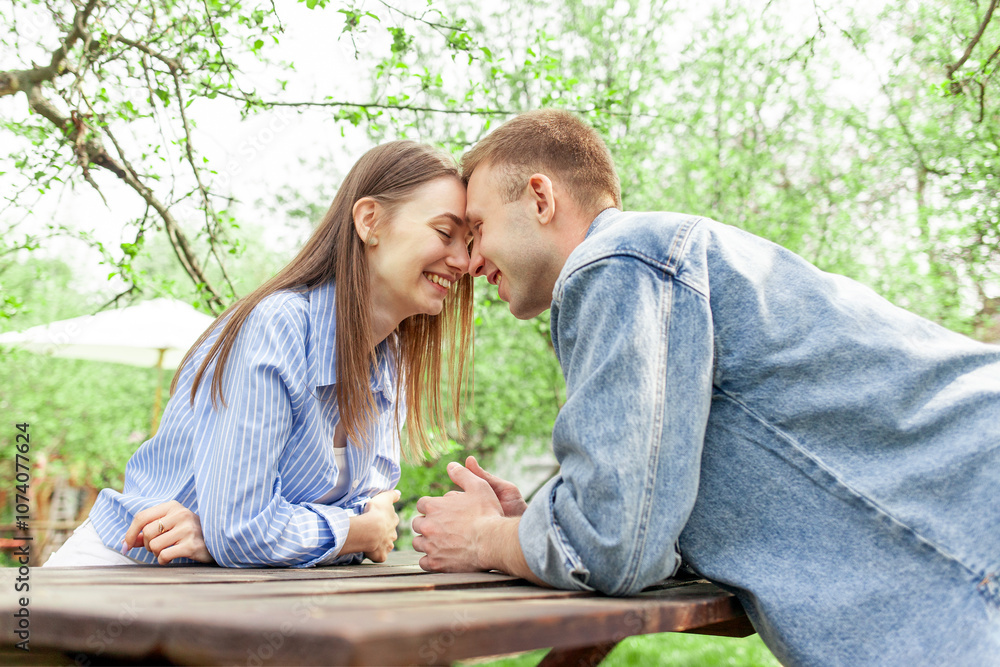 young couple in love in denim clothes sitting in the garden at a wooden table and talking in the summer, guy and girl on a date talking and flirting in the park young couple in love in denim clothes sitting in the garden at a wooden table and talking in the summer, guy and girl on a date talking and flirting in the park