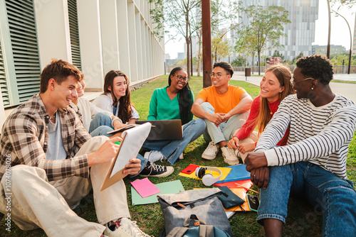 diverse group of happy university students sitting together on green campus lawn while studying reading and discussing with notebooks laptop and backpack during outdoor learning session