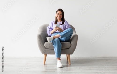 Fotografie Beautiful young woman sitting in armchair, looking at mobile phone screen, using new cool application against white studio wall, full length