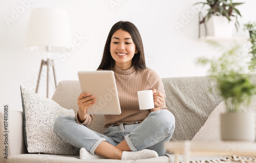 Cheerful girl reading e-book on digital tablet, drinking morning coffee on sofa photo