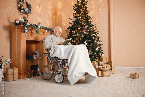 Old man on a wheelchair sitting near Christmas tree and reading a book