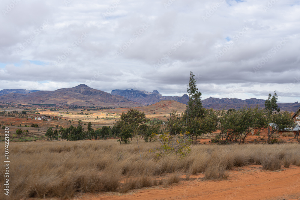 Fototapeta premium Expansive farmland with patches of green and brown fields under a cloudy sky. Rural landscape, Isalo National Park, Madagascar.