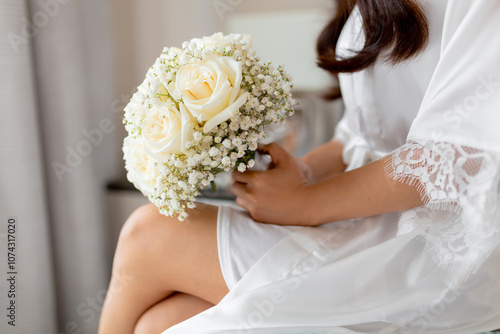 Wedding bouquet of white roses and gypsophila in the hands of the bride, close-up. The bride is sitting in a room in a white silk robe and holding a wedding bouquet before the wedding ceremony photo