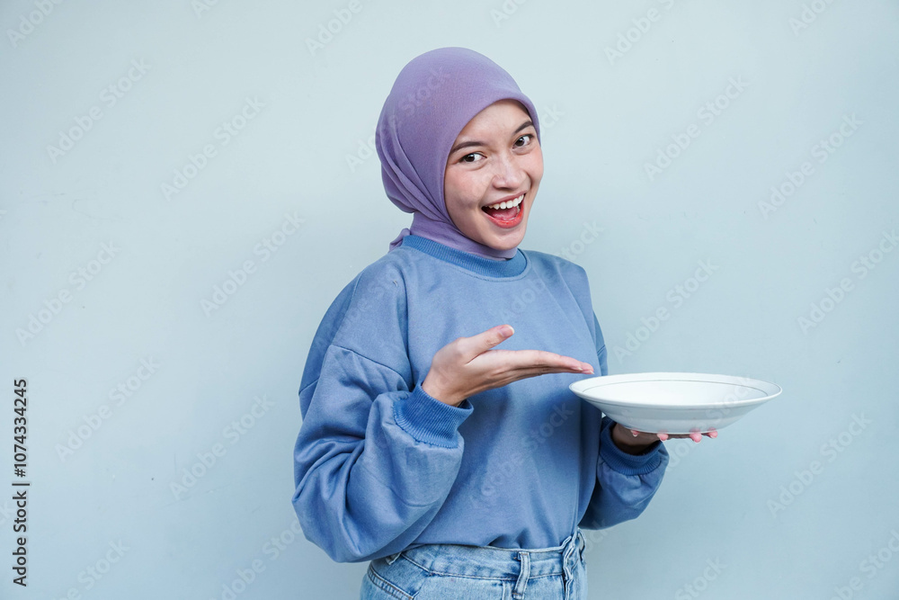 A smiling Asian Muslim woman is fasting and hungry and holding and pointing to a plate. A smiling Asian Muslim woman is fasting and hungry and holding and pointing to a plate.