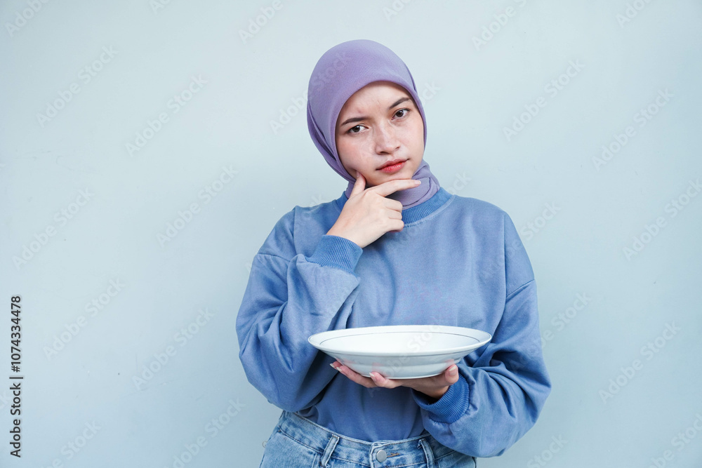 Asian Muslim woman is fasting and hungry and holding cutlery while looking aside thinking about what to eat with empty plate. Asian Muslim woman is fasting and hungry and holding cutlery while looking aside thinking about what to eat with empty plate.