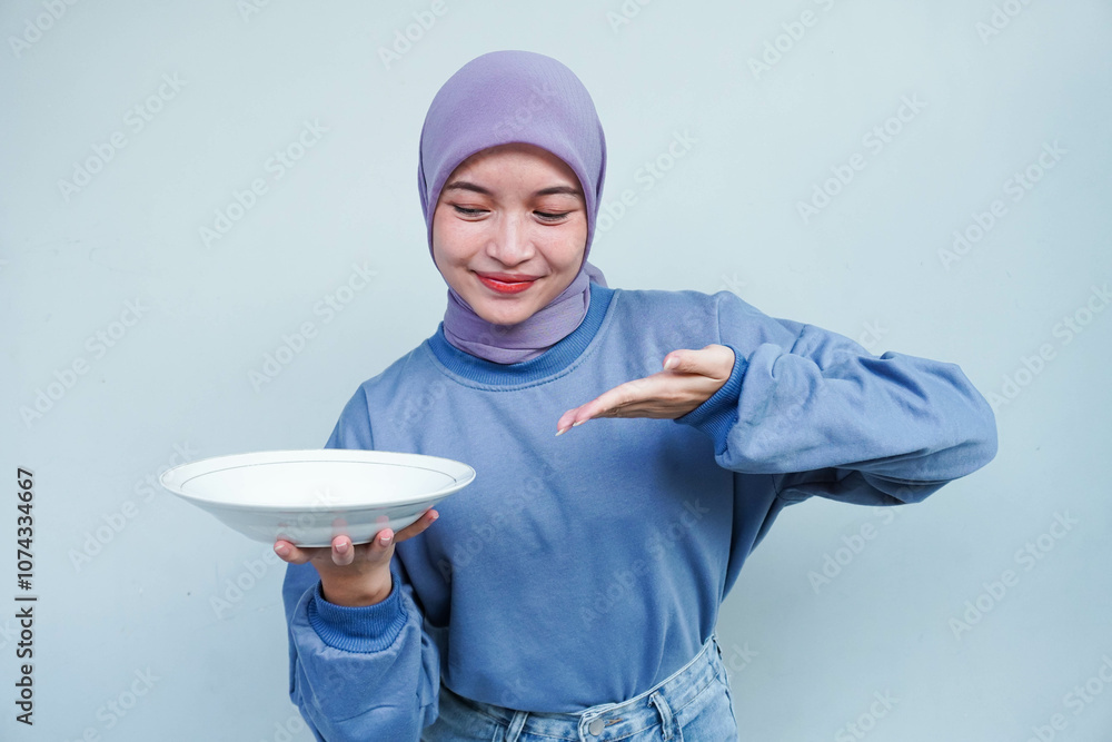 A smiling Asian Muslim woman is fasting and hungry and holding and pointing to a plate. A smiling Asian Muslim woman is fasting and hungry and holding and pointing to a plate.