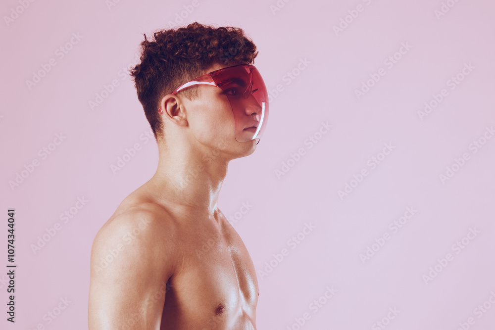 Stylish young man with curly hair wearing bold red visor sunglasses, showcasing a modern fashion statement against a soft pink background. Stylish young man with curly hair wearing bold red visor sunglasses, showcasing a modern fashion statement against a soft pink background.