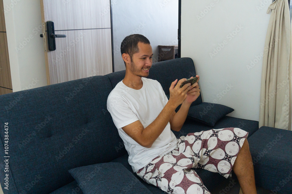 Obraz premium Young asian men wearing white shitr is holding smart phone with smile on sofa.