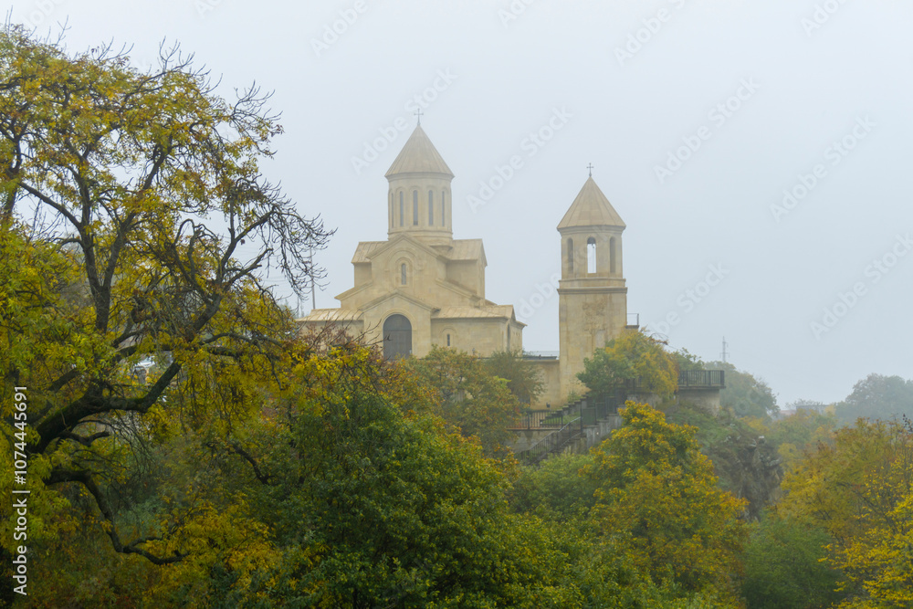 Naklejka premium Church of St. Ekvtime and bell tower in Kiketi village on a foggy morning. Trees with autumn leaves, bushes in the foreground