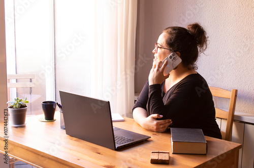 Young, white, smiling woman working from home in her private office. With a computer, a book, pens and a cell phone. Wooden table, black clothes.