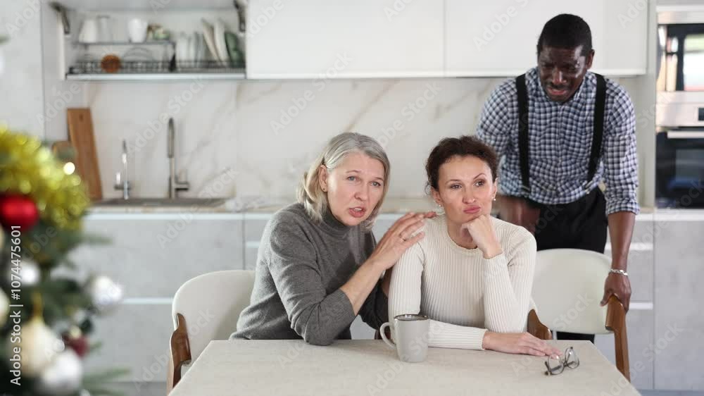 Elderly woman calms adult woman during family quarrel with man in kitchen Elderly woman calms adult woman during family quarrel with man in kitchen