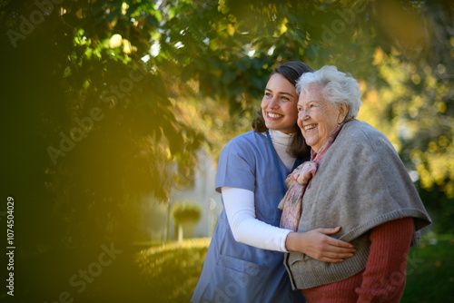 Portrait of nurse and elderly woman on walk in the park during warm autumn day. Young caregiver spending time with senior patient.
