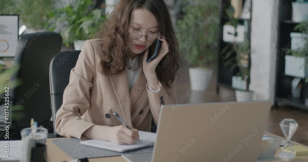 Asian office worker speaking on mobile phone writing information in notebook and using laptop computer. Business and communication concept. Asian office worker speaking on mobile phone writing information in notebook and using laptop computer. Business and communication concept.