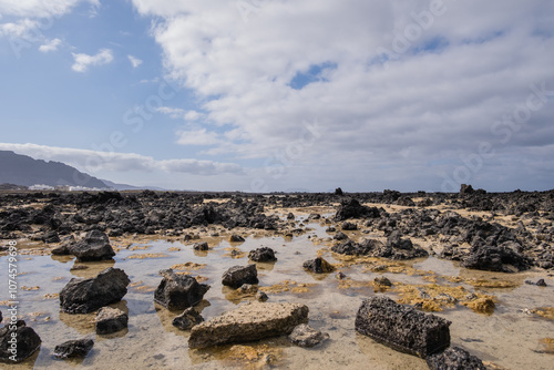 White sand beach with black rocks of volcanic origin. Mountains in the background and sky with big white clouds. Caletón Blanco, Lanzarote, Canary Islands, Spain