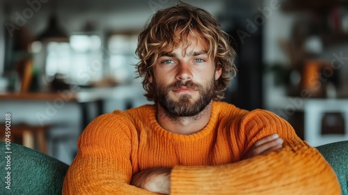 This image shows a man with curly hair sitting snugly, wearing an orange sweater and projecting a calm demeanor, set in a cozy, modern living space filled with light. photo