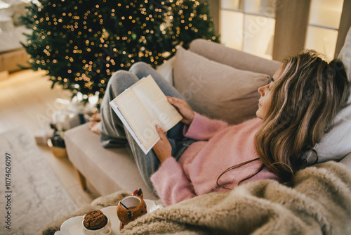 Young woman relaxing, reading book and drinking cocoa in a cozy living room near Christmas tree.