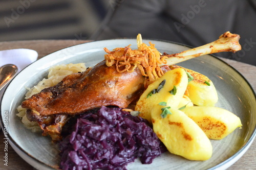 close view of roasted goose legs, red and white cabbage, potato dumplings, served food on a plate in a restaurant, wooden table, St. Martin's goose, traditional Czech holiday food photo