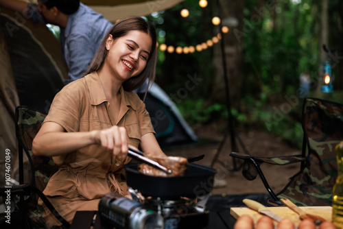 asian woman grilling meat on a portable gas stove while camping in nature park at evening time. Travel, adventure and vacation concept