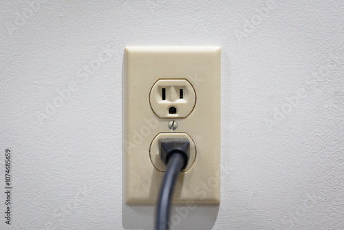 Wall outlet on a white wall with one plug connected. North American outlet with beige outlet plate
